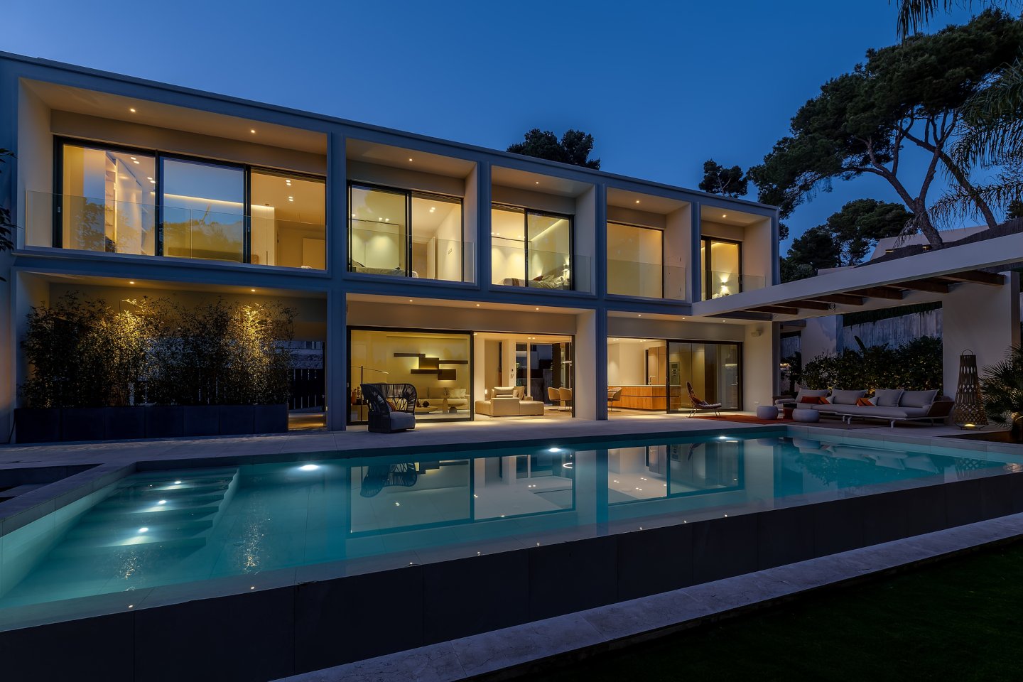 Elegant pool area at night in a contemporary beachside villa in Bendinat, Mallorca, featuring a beautifully lit infinity pool surrounded by sleek modern design elements. The ambient lighting creates a serene and luxurious atmosphere, highlighting the villa's architectural beauty and offering a perfect setting for evening relaxation and entertainment.