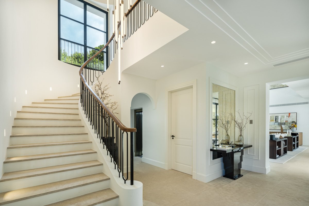 The grand entrance of an elegant villa in Cascada de Camoján, Marbella, with a sleek, modern design featuring large wooden doors, elegant stone accents, and a beautifully landscaped pathway. The entrance is framed by lush greenery and tall palm trees, setting a sophisticated tone for the luxurious home on the Golden Mile.