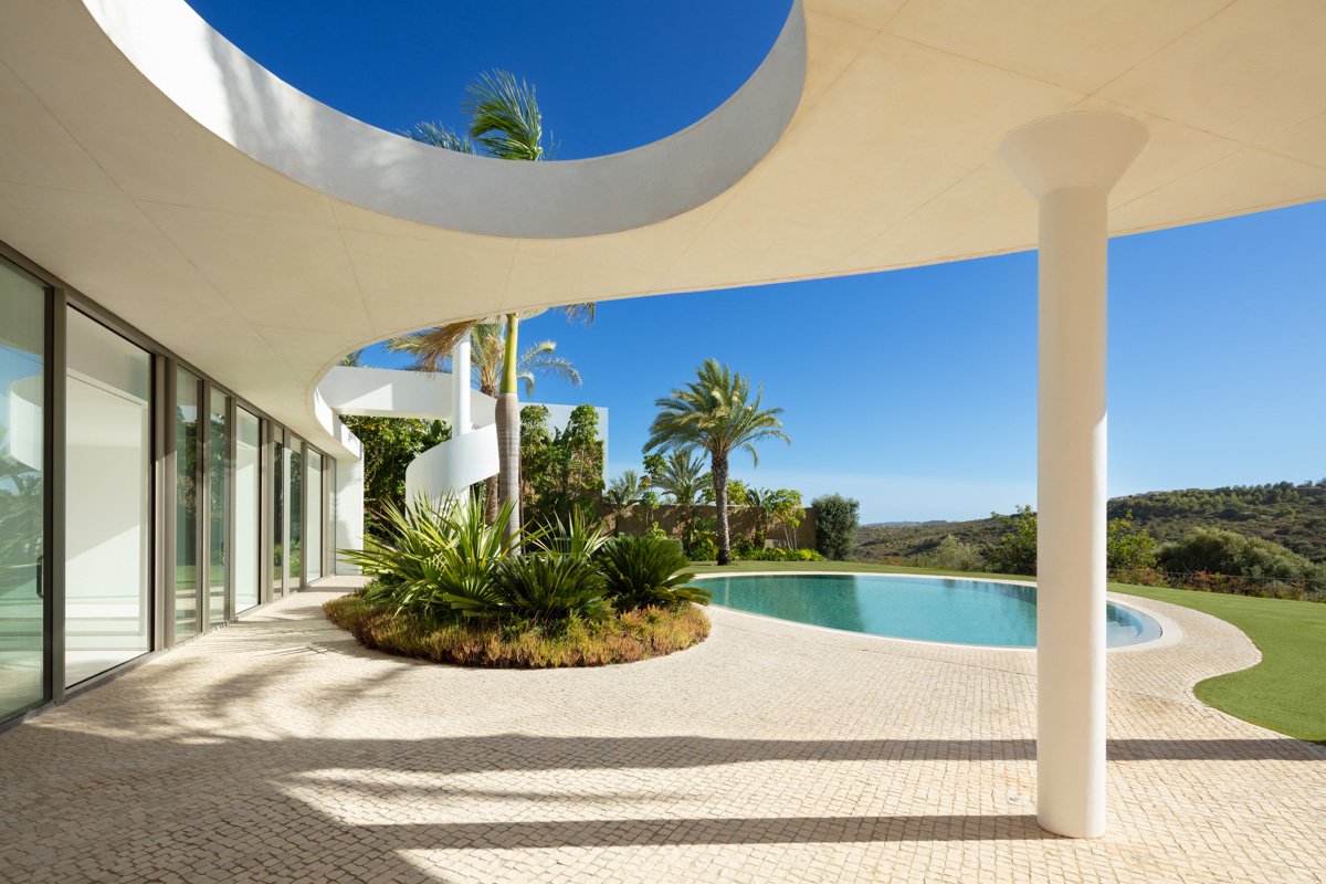 A breathtaking view of a futuristic villa in Finca Cortesin, Marbella, showcasing cutting-edge architecture with sleek lines and expansive glass windows. The villa is surrounded by lush landscaping and offers stunning panoramic views of the golf course and the natural beauty of the area.