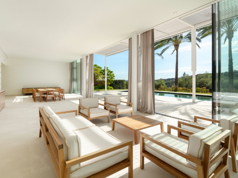 A spacious and elegant living room with modern, comfortable seating, a large flat-screen TV, and a striking contemporary design. Floor-to-ceiling windows flood the room with natural light, offering stunning views of the surrounding landscape. Neutral tones and refined decor create a sophisticated yet welcoming atmosphere.