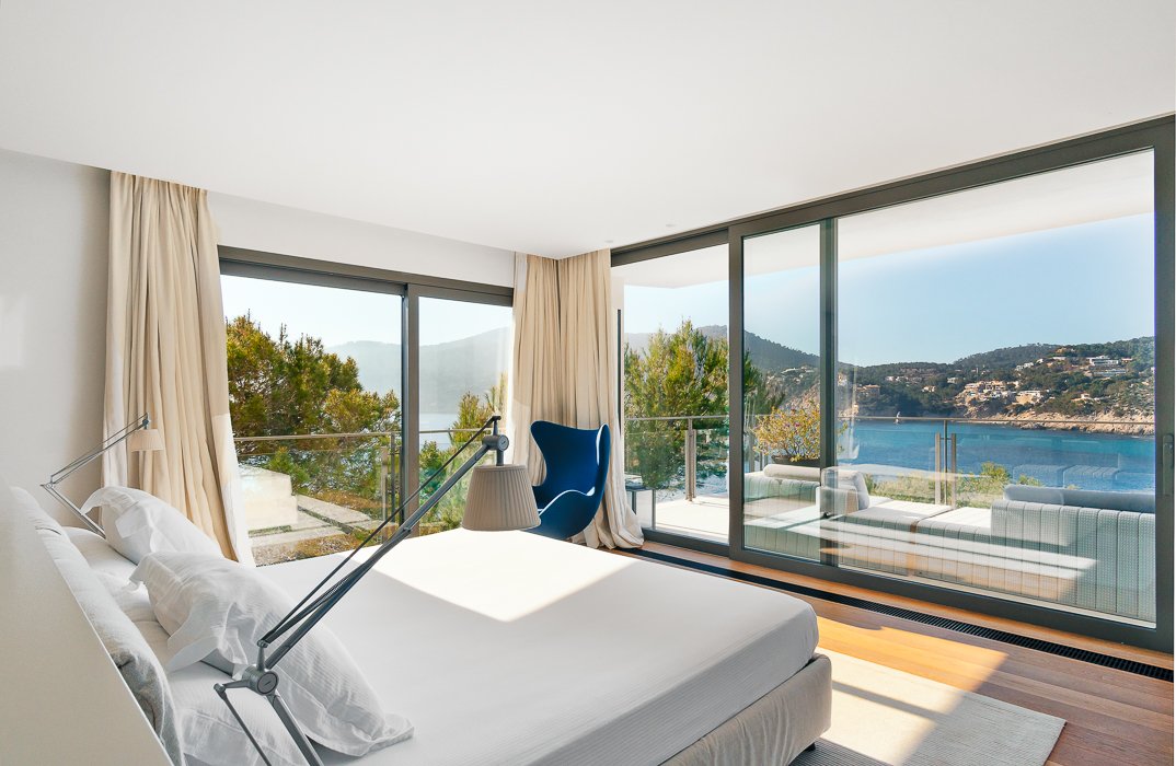 Elegant bedroom in a seafront luxury villa in Camp de Mar, Mallorca, featuring modern furnishings, soft lighting, and large glass doors leading to a private balcony. The balcony offers breathtaking views of the Mediterranean Sea, creating a peaceful and luxurious retreat.